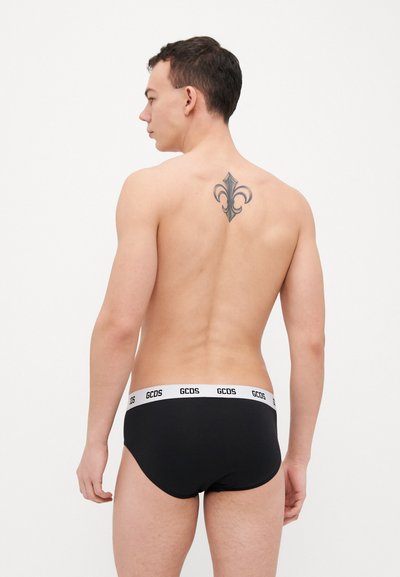 Black cotton briefs with a white logo waistband, showing a fitted design. The model has a tattoo on the upper back.