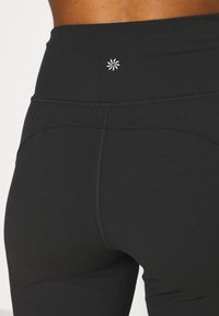 Leggings neri con un alto punto vita e un piccolo logo bianco sul retro. Il tessuto sembra liscio ed elastico con cuciture piatte.