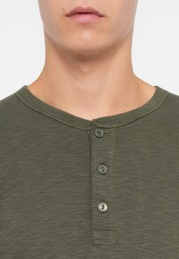 Maslinasto zelena henley majica izrađena od mekanog materijala, s okruglim izrezom, tri sive dugmadi i teksturiranim završetkom.