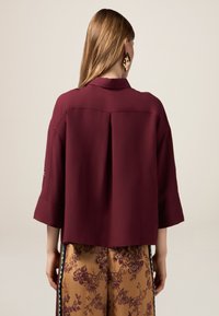 Burgundy lös passande blus med trekvartslånga ärmar och detaljer med veck på baksidan, kombinerad med beige byxor med blommönster.