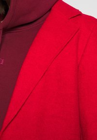 Rode wolmix blazer met scherpe revers, gestructureerde stof en een bordeauxrode hoodie die gedeeltelijk zichtbaar is eronder.