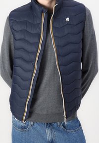 Man med en marinblå vadderad väst över en grå stickad tröja och blå denimjeans, med händerna bakom ryggen och dragkedjan delvis öppen.