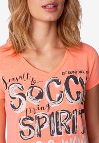 Camiseta de manga corta color coral con cuello en V, que presenta un texto gráfico en blanco y negro "Seasalt & Soccy Living Spirit", adornada con lentejuelas.