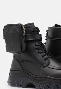 Bottes de combat en cuir noir avec des semelles épaisses et robustes et une petite pochette zippée attachée près du haut.