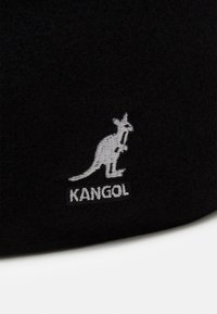 Sombrero de lana negro con un logotipo de canguro bordado en blanco y el texto "KANGOL" debajo. La tela parece suave y texturizada.