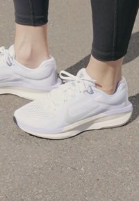 Bijele sportske tenisice s mrežastim gornjim dijelom, zaobljenim vrhom i suptilnim sivim naglascima. Lagana potplat s teksturiranim donjim dijelom za prianjanje. Uparene s crnim tajicama.
