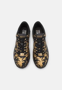 Baskets noires avec un motif baroque doré, dotées de lacets noirs, semelles rembourrées et une semelle intérieure de marque. Détail accent doré sur la languette.