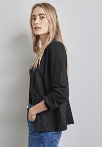 Černý blazer s mírně strukturovanou tkaninou, na jediné knoflíkové zapínání a s ohrnutými rukávy. Nosený přes tmavý top a kombinovaný s modrými džíny.