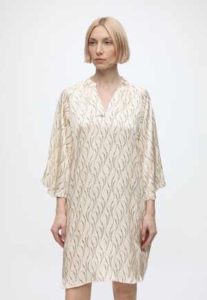 Kvinde med kort blondt hår iført beige knælængde kjole med sort bølget linje- og prikkemønster samt trekvart pufærmer.