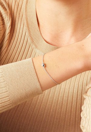 Poignet portant un délicat bracelet chaîne en argent avec un petit pendentif en forme de cœur, assorti à un pull beige à manches longues côtelé.