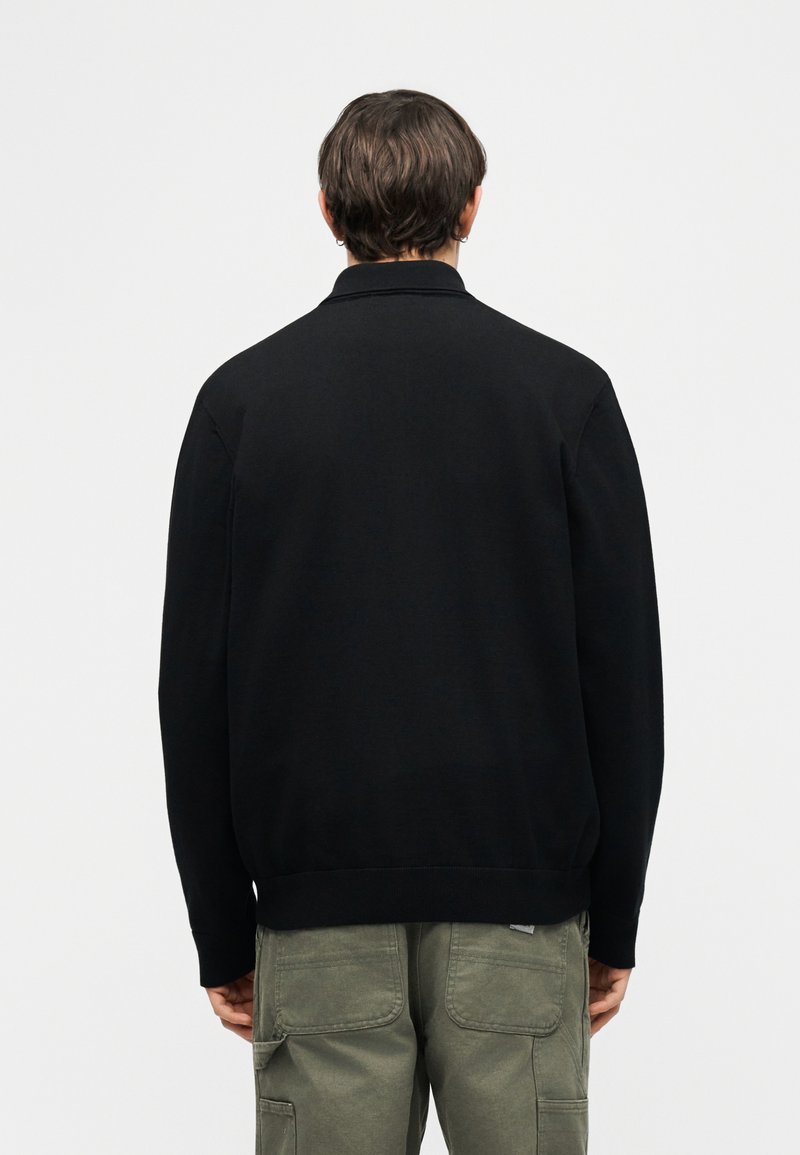 Junger Mann, der ein schlichtes schwarzes Langarmshirt und olivgrüne Cargohosen trägt, von hinten vor weißem Hintergrund gesehen.