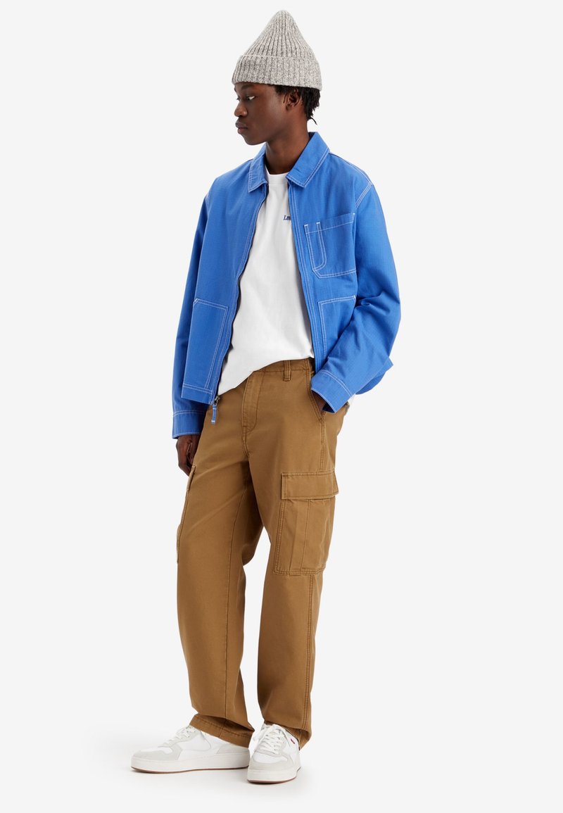 Chaqueta de trabajo azul con costuras blancas, camiseta blanca, pantalones cargo marrones con bolsillos laterales y zapatillas blancas. Gorro de lana gris.