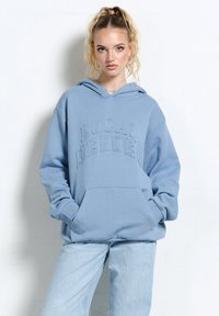 Młoda kobieta o kręconych blond włosach związanych w wysoki kucyk, ubrana w niebieską bluzę z kapturem "Local Heroes" i jasnoniebieskie dżinsy, stoi na tle białej ściany.