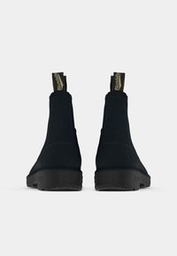 Para ciemnoniebieskich zamszowych botków za kostkę z czarnymi podeszwami i markowymi pętelkami, widocznych z tyłu na białym tle.