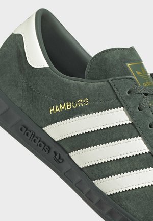 Zelený semišový teniska Adidas s bílými pruhy, černou podrážkou, zlatým nápisem „HAMBURG“ a logem Adidas na jazyku a boku.