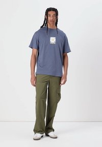 Tricou albastru din bumbac cu un imprimeu grafic în centru, asociat cu pantaloni cargo verzi și adidași albi cu accente bej.