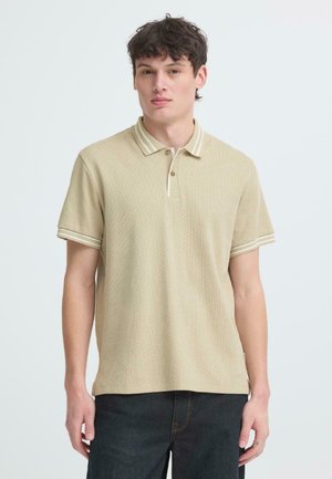 Jeune homme aux cheveux courts et foncés, vêtu d'un polo beige texturé à manches courtes et d'un pantalon foncé, debout devant un fond clair uni.