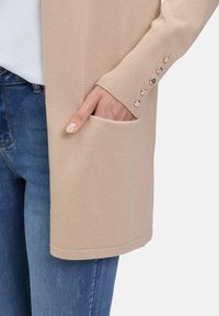 Beige gebreide cardigan met een geribbelde manchet, voorzien van vijf decoratieve knopen en een zijzak. Gecombineerd met blauwe spijkerbroek.