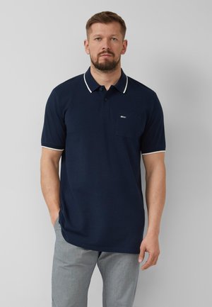 Man draagt een navyblauw poloshirt met korte mouwen en witte bies aan kraag en mouwen, gecombineerd met lichtgrijze broek, staand tegen een grijze achtergrond.