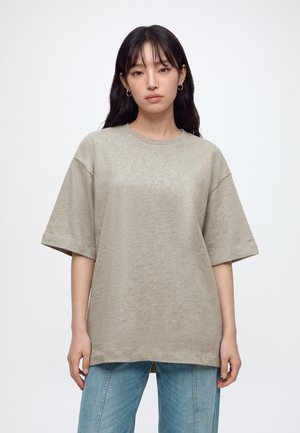 Donna con lunghi capelli scuri che indossa una maglietta beige larga e jeans azzurri chiari, in piedi davanti a uno sfondo bianco semplice.