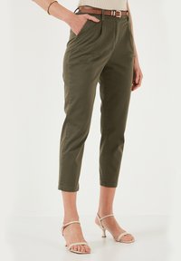 Pantaloni in cotone verde oliva con una vestibilità affusolata, caratterizzati da una piega frontale, tasche laterali e una cintura marrone. Indossati con sandali chiari.