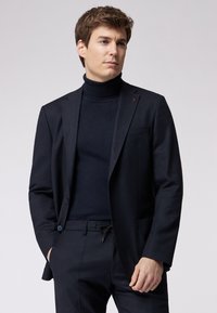Marineblazer mit glatter Textur, ausgestattet mit einem Ein-Knopf-Verschluss und einer Vorderasche. Über einem passenden Rollkragenpullover getragen, der einen abgestimmten Look zeigt.
