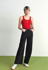 Raudonas tankų marškinėlis su logo, juodos aukštu liemeniu korėtos plonos plačios kelnės ir baltos sportbačiai su oranžiniais padais. Lygus fonas.