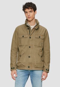 Khaki jas met een opstaande kraag, vier voorzakken, zwarte knopen en lichte stikdetails, gecombineerd met blauwe jeans.