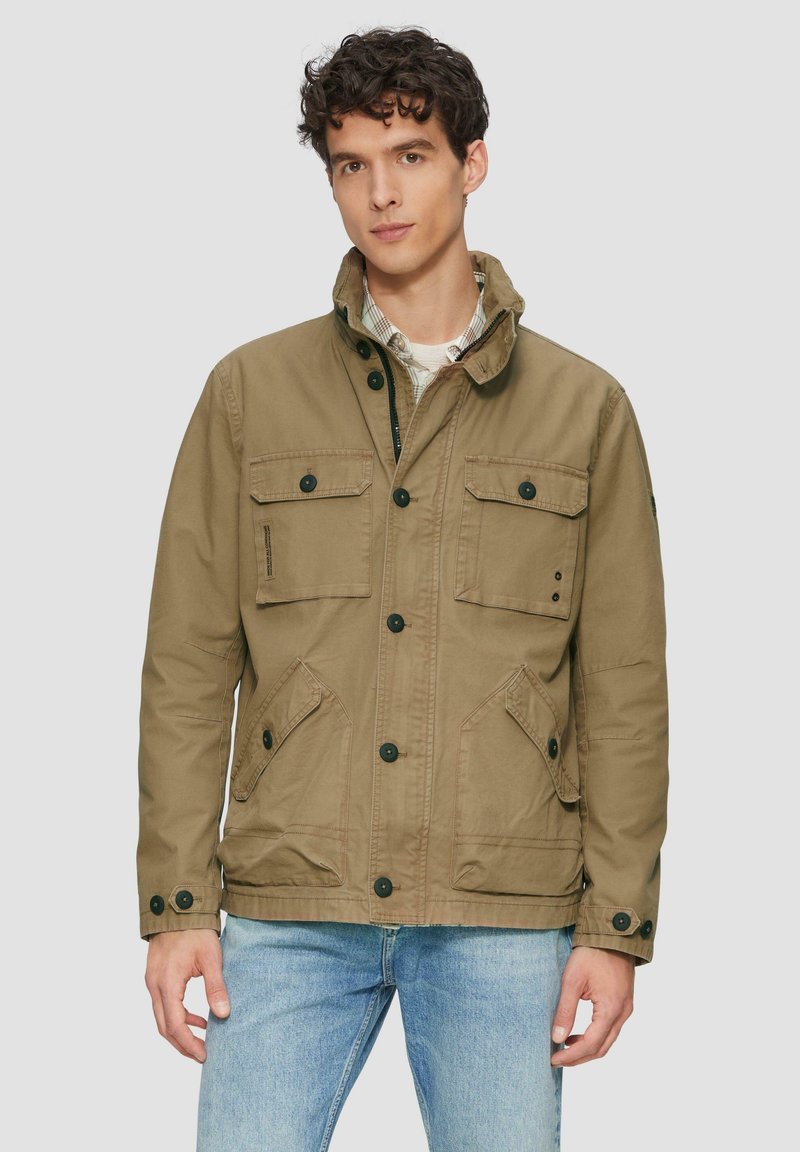Khaki jas met een opstaande kraag, vier voorzakken, zwarte knopen en lichte stikdetails, gecombineerd met blauwe jeans.