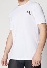 Camiseta blanca de manga corta hecha de tela suave, con un logo negro de "Under Armour" en el lado izquierdo del pecho. Cuello redondo estándar.