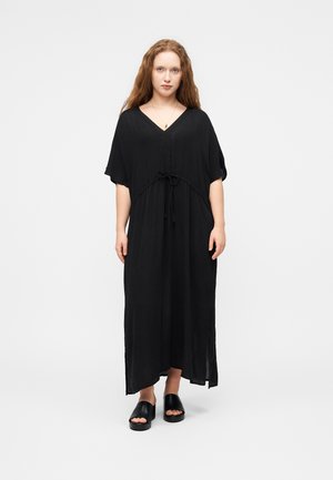 Kobieta o długich rudych włosach, ubrana w czarną luźną sukienkę maxi z dekoltem w serek i krótkimi rękawami oraz czarne sandały wsuwane, stojąca na jednolitym tle.