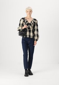 Črno-bela karirana bluzica z nagubanimi izrezi, kombinirana z temno modrimi jeans hlačami in črnimi gležnjarji; model drži črno torbico za ramo.