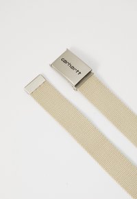 Bežový pletený opasok s kovovou prackou s logom "Carhartt". Pás má textúrovaný vzor a končí kovovými štvorcovými špičkami.