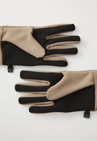 Beige og svarte hansker med teksturert overflate, fleksibelt materiale og detaljer på fingertuppene, samt en justerbar stropp for sikker passform.