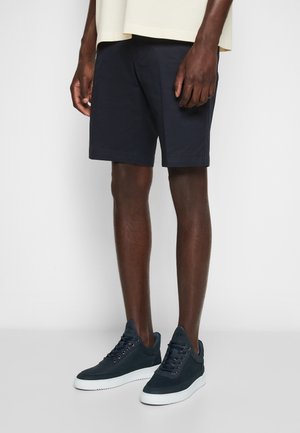 Homme portant un short bleu marine foncé et des baskets noires avec des semelles blanches, debout devant un fond blanc uni.
