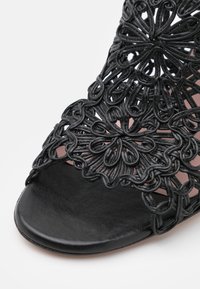 Gros plan sur une sandale noire à bout ouvert avec des brides florales complexes en boucles et tressées sur le dessus du pied.