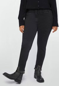 Pantalones de mezclilla negros con una cinturilla elástica, con detalles de tachuelas en los tobillos. Llevados con una camisa negra abotonada y botas negras.