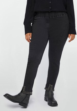Pantaloni din denim negru cu talie elastică, ornati cu accente cu ținte la glezne. Purtați cu o cămașă neagră cu butoni și cizme negre.