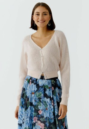 Lächelnde Frau mit einem hellrosa, flauschigen, geknöpften Cardigan und einem taillenhohen, marineblauen Rock mit großem blauem und pinkem Blumenmuster.