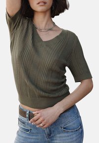 Pull à manches courtes vert olive avec un col en V, présentant une texture côtelée et des motifs en maille ouverte, associé à un jean bleu clair et une ceinture marron.