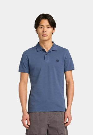 Uomo che indossa una polo blu a maniche corte con un piccolo logo sul petto e pantaloncini grigi, in piedi davanti a uno sfondo semplice.