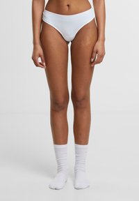Witte katoenen slip met hoge beenuitsnijding en elastische tailleband, gecombineerd met witte geribde crew sokken op blote voeten. Glad textuur, minimalistisch design.
