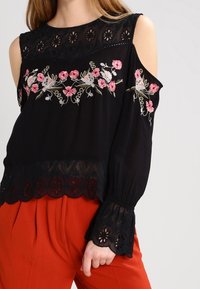 Svart kall-axel topp med blommig broderi, spetsdetaljer och scallopad nederkant. Långa ärmar med blommiga accenter vid ärmsluten.