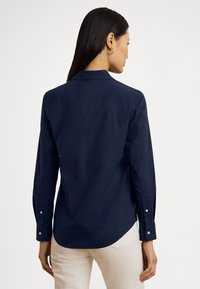 Temno modra dolgo rokavna srajca iz gladkega blaga z nazobčanim ovratnikom, nazaj šivom in zapognjenimi manšetami na gumbe. Preprost, minimalističen dizajn.
