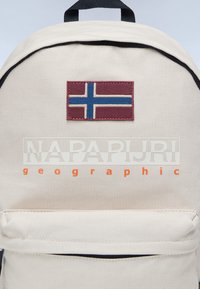 Bežový plátěný batoh s vínovým nápletem, na kterém je modro-bílá norská vlajka. Obsahuje význačné logo značky v bílé a oranžové barvě.