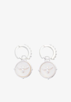 Boucles d'oreilles créoles en argent avec pendentifs ronds blancs arborant un motif d'abeille en relief sur fond blanc lisse.