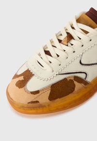 Nærbilde av beige og brune sneakers med hvite lisser, camouflagemønstrede tå og gjennomsiktig oransje såle på hvit bakgrunn.