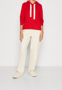 Sudadera roja con cordones blancos, combinada con pantalones de pierna ancha color crema y zapatillas beige. Tejido suave con un ajuste relajado.