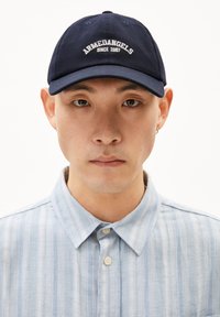 Marineblauwe baseballpet met witte geborduurde tekst "ARMEDANGELS SINCE 2007" aan de voorkant, gedragen met een lichtblauwe gestreepte overhemden.