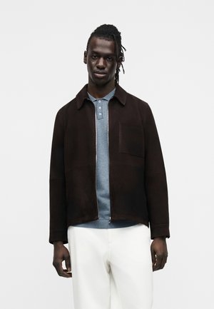 Braune Wildlederjacke mit Kragen, frontalem Reißverschluss und einer Brusttasche, getragen über einem hellblauen Polohemd mit Struktur und weißen Hosen.